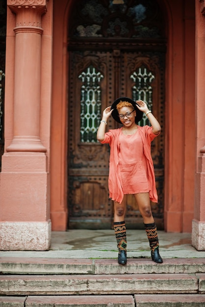 Une femme africaine aux cheveux rouges porte des lunettes, une robe orange et un chapeau noir posant en plein air.