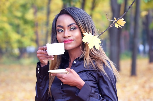 Femme africaine de boire du café dans une tasse blanche sur le fond des feuilles d'automne dans le parc