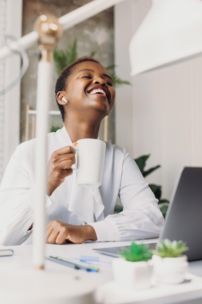 Femme africaine buvant du thé pendant la pause déjeuner et utilisant des écouteurs pour parler à ses amis avec qui