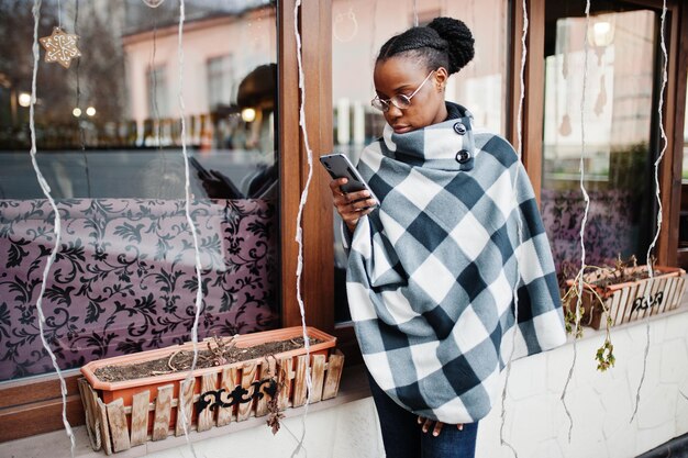 Femme africaine en cape à carreaux et lunettes posées en plein air et regarder le téléphone mobile