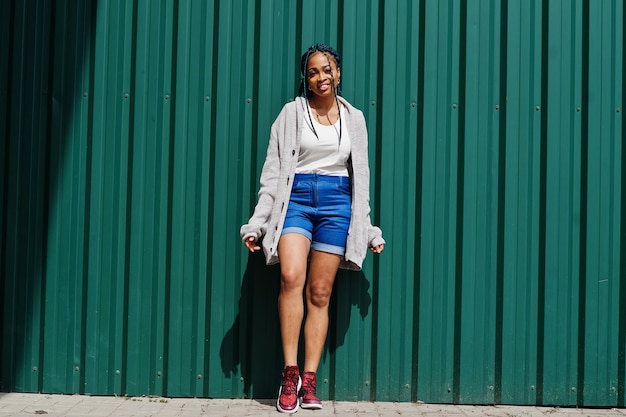 Femme africaine avec des cheveux dreads en short jeans posé contre un mur d'acier vert