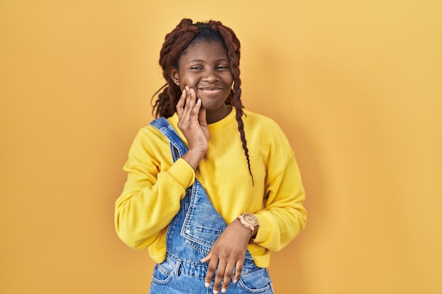 Femme africaine debout sur fond jaune touchant la bouche avec la main avec une expression douloureuse à cause d'un mal de dents ou d'une maladie dentaire sur les dents. dentiste