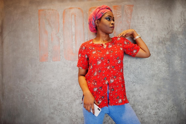 Femme africaine élégante en chemise rouge et chapeau posé contre le mur avec signe de rock