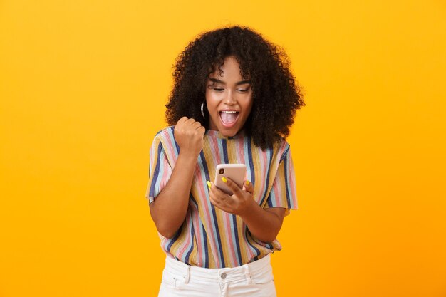 Femme africaine émotionnelle posant isolée sur un espace jaune à l'aide de téléphone mobile faire le geste gagnant.