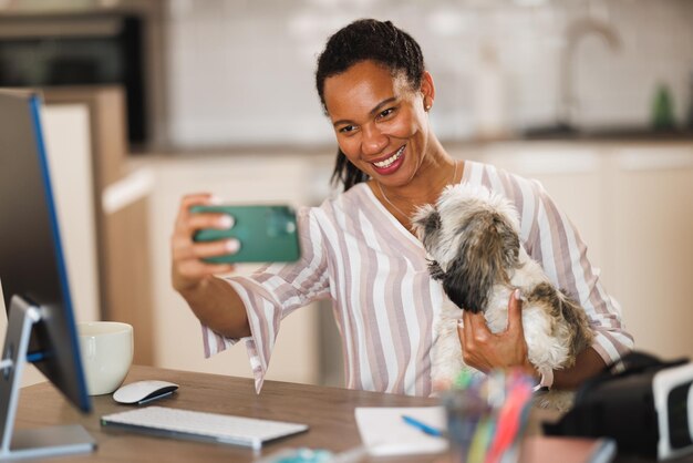 Femme africaine faisant du selfie et s'amusant avec son chien de compagnie tout en travaillant à son bureau à domicile.