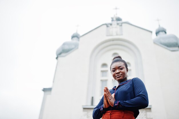 Femme africaine en pantalon orange et chemise bleue posée contre une grande église Foi et croire en Dieu