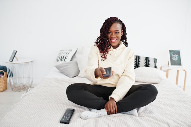 Femme africaine regardant la télévision à la maison et tenant une télécommande sur le lit.