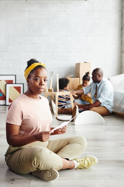 Femme africaine sérieuse avec du papier assis sur le sol du salon contre un jeune homme et deux enfants assemblant une chaise ensemble