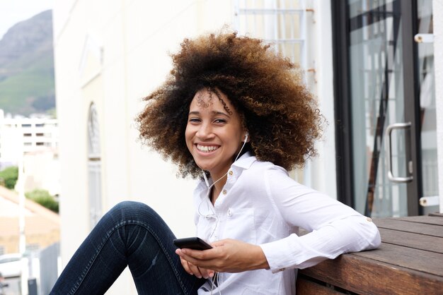 Femme africaine souriante, assis à l&#39;extérieur avec un téléphone intelligent et des écouteurs