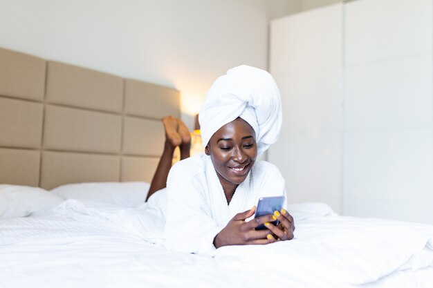 Femme africaine souriante dans un peignoir allongé sur un lit avec téléphone portable, textos et souriant assis sur un lit.