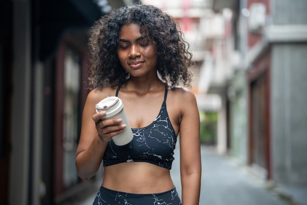 Femme africaine sport en forme saine et ferme mince boire de l'eau de la tasse sur la main course athlétique portant dans les vêtements de sport à la ville et à la ville Motivation de remise en forme et de sport Concept de coureur