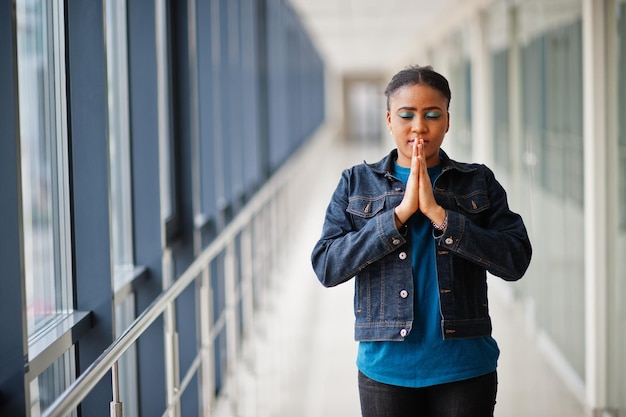 Une femme africaine en veste de jeans prie à l'intérieur.