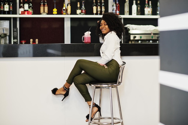 Une femme afro-américaine d'affaires pure aux cheveux afro porte un chemisier blanc et un pantalon vert assis sur le comptoir du bar et boit un cocktail rose