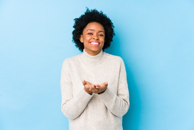 Femme afro-américaine d'âge moyen contre un bleu isolé tenant quelque chose avec des palmiers, offrant à la caméra.