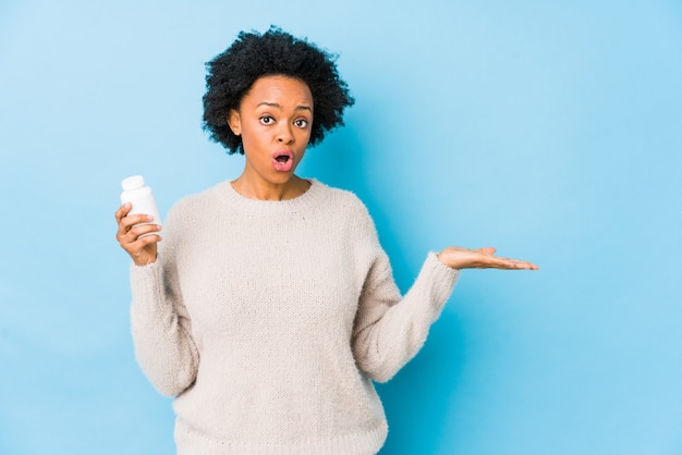 Femme afro-américaine d'âge moyen tenant une bouteille de vitamine impressionné tenant copie espace sur la paume.