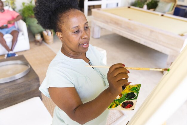 Photo une femme afro-américaine âgée et heureuse qui tient une palette et peint une toile sur un chevalet. style de vie, retraite, créativité, loisirs, art et vie domestique, inchangés.