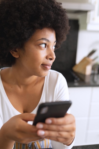 Femme afro-américaine assise dans la cuisine en utilisant un smartphone et en pensant