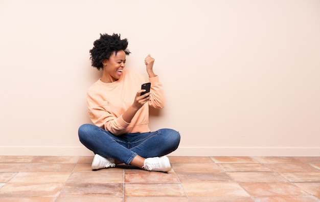 Femme afro-américaine assise sur le sol avec téléphone en position de victoire