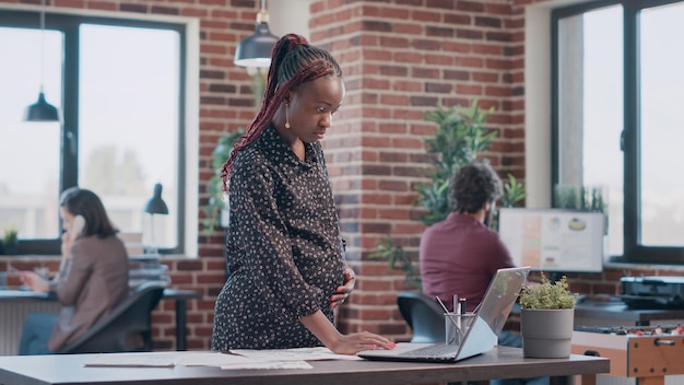 Une femme afro-américaine attend un enfant et travaille sur un projet d'entreprise avec des documents. Employée enceinte analysant des fichiers pour concevoir et planifier une stratégie marketing pour le développement au bureau.