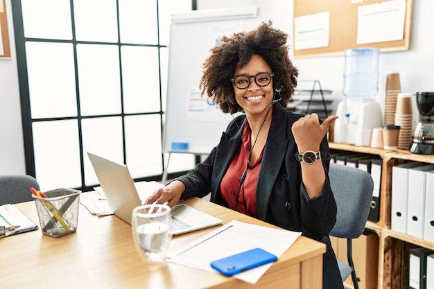 Femme afro-américaine aux cheveux afro travaillant au bureau portant un casque d'opérateur pointant vers l'arrière derrière avec la main et les pouces vers le haut, souriant confiant