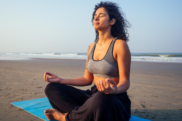 Femme afro-américaine aux cheveux bouclés et aux yeux fermés respirant profondément et se calmant sur la plage vide du matin après surya namaskar