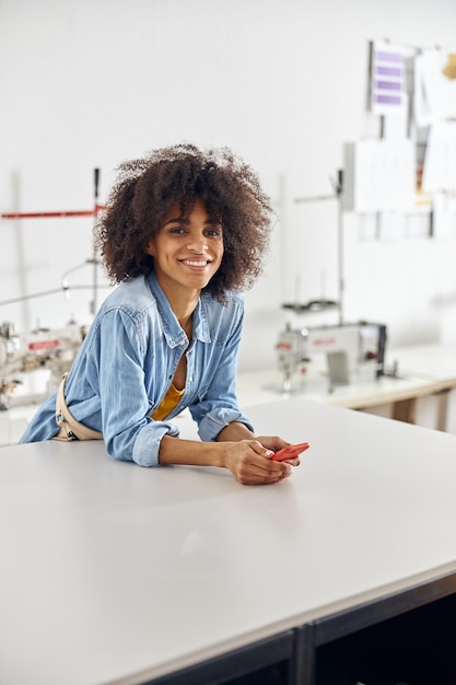 Une femme afro-américaine aux cheveux bouclés en chemise en jean se penche sur une table de coupe en atelier