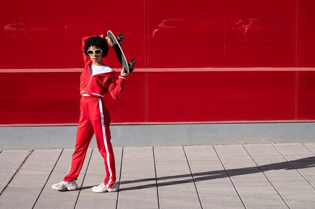 Photo femme afro-américaine aux cheveux courts et en vêtements de sport avec un skateboard sur fond rouge en ville