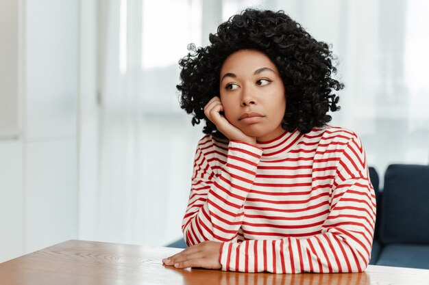 Photo femme afro-américaine bouleversée portant des vêtements décontractés regardant loin, assise au bureau dans le salon