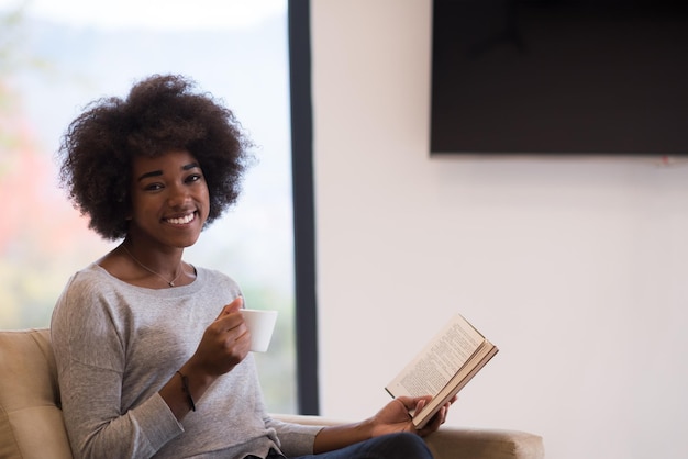 femme afro-américaine buvant une tasse de café lisant un livre à la cheminée. Jeune fille noire avec une boisson chaude relaxante chauffant l'échauffement. automne à la maison.