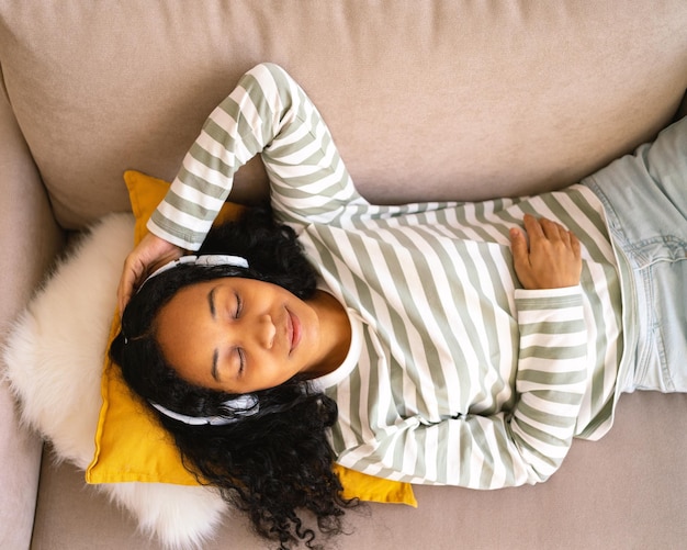 Photo femme afro-américaine calme allongée sur un canapé en écoutant de la musique dans un casque en profitant de la lecture audio