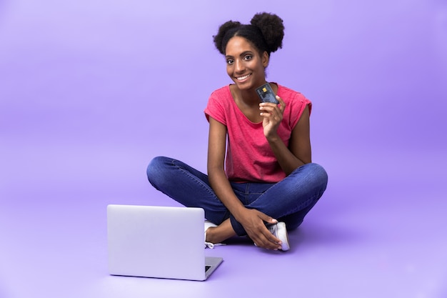 Photo femme afro-américaine candide souriant et utilisant un ordinateur portable d'argent, alors qu'il était assis sur le sol avec les jambes croisées, isolé