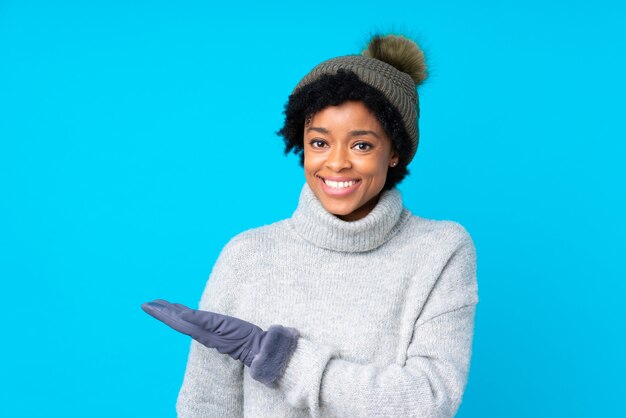 Femme afro-américaine avec chapeau d'hiver sur un mur bleu isolé