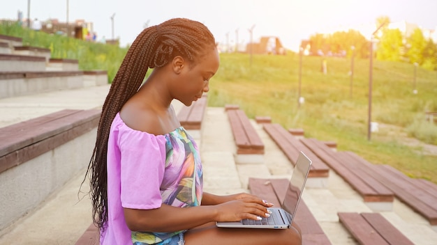 Une femme afro-américaine concentrée en blouse de couleur élégante travaille sur un ordinateur portable moderne assis sur un banc dans un parc d'été verdoyant par beau temps