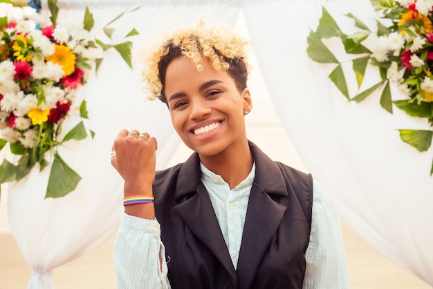 Femme afro-américaine en costume de marié noir attendant sa mariée près de l'arche florale de mariage sur la plage tropicale portant un bracelet arc-en-ciel