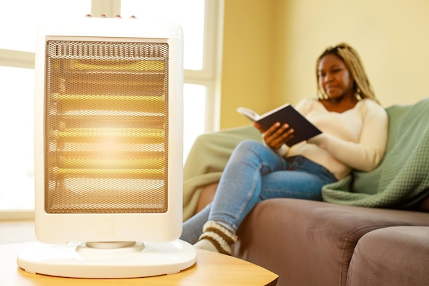 Femme afro-américaine en couverture bleue lisant un livre près d'un radiateur électrique à la maison