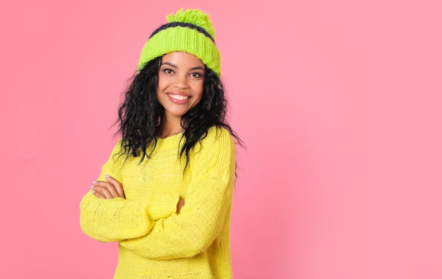 Une femme afro-américaine dans un pull jaune vif et un chapeau vert à pompon se tient dans un demi-profil, les bras croisés, regardant la caméra avec un grand sourire