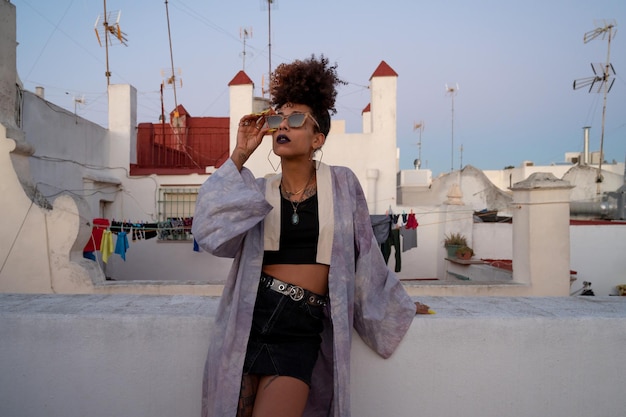 Femme afro-américaine dans des vêtements élégants ajustant des lunettes de soleil et s'appuyant sur la barrière contre les maisons tout en se relaxant sur la terrasse le soir à Cadix en Espagne