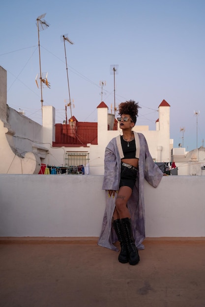 Femme afro-américaine dans des vêtements élégants avec des lunettes de soleil et s'appuyant sur la barrière contre les maisons sur la terrasse en soirée CadizSpain