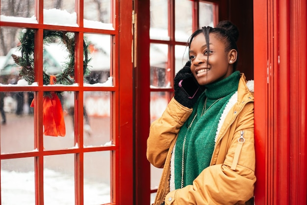 Femme afro-américaine debout à l'extérieur de la rue près du marché équitable de l'arbre de Noël, tenir le téléphone parlant