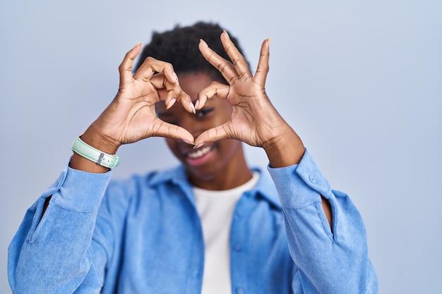 Femme afro-américaine debout sur fond bleu faisant la forme de coeur avec la main et les doigts souriant regardant à travers le signe