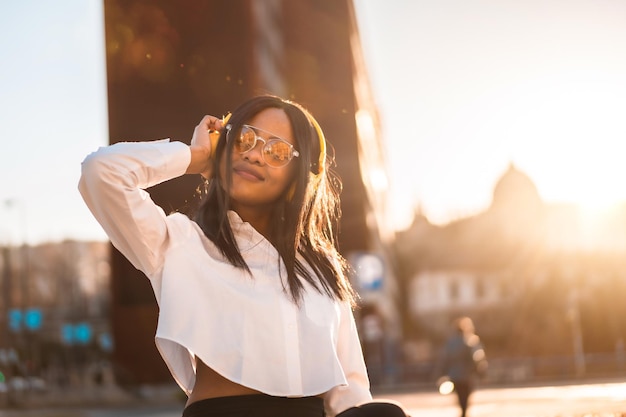 Femme afro-américaine écoutant de la musique avec des écouteurs au coucher du soleil dans la musique de la ville en ligne et dansant en souriant