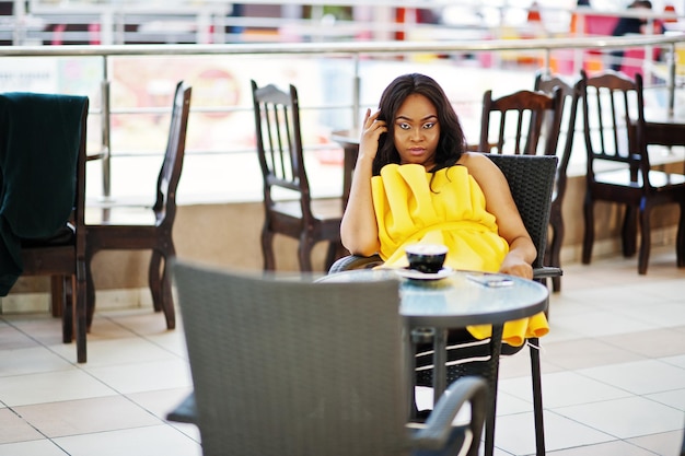 Femme afro-américaine élégante à la robe jaune posée au café avec une tasse de cappuccino