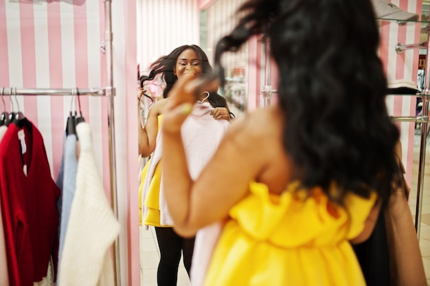 Photo femme afro-américaine élégante à la robe jaune posée dans la boutique rose et choisit des vêtements contre le miroir