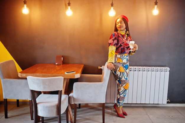 Femme afro-américaine enthousiaste en tenue colorée à la mode avec béret rouge se relaxant dans un café confortable avec une tasse de latte chaud dans les mains