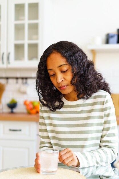 Photo une femme afro-américaine épuisée attend que la pilule se dissolve dans un verre d'eau dans la cuisine.