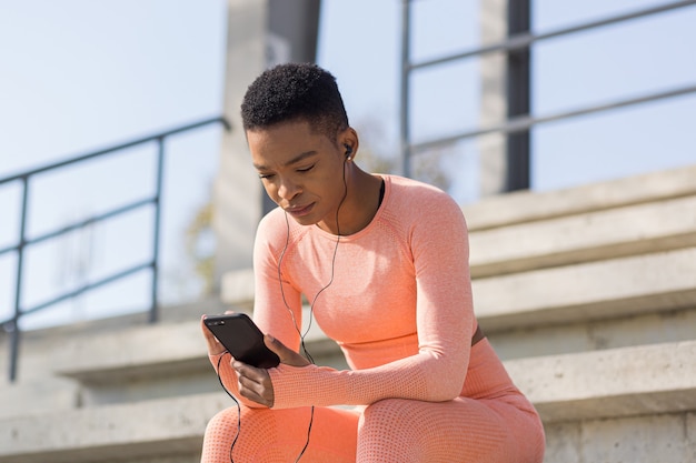 Femme afro-américaine fatiguée après l'entraînement en écoutant de la musique et un podcast audio, après avoir fait du jogging et s'être entraîné au stade le matin