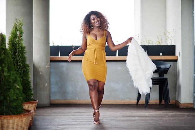 Une femme afro-américaine glamour en robe jaune et en cape de laine blanche pose sur la terrasse