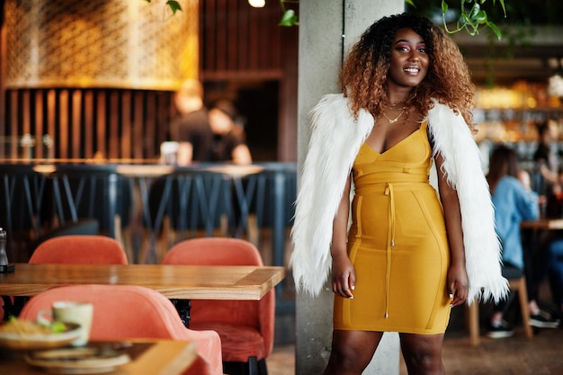 Femme afro-américaine glamour en robe jaune et cape de laine blanche posée au restaurant près de la colonne