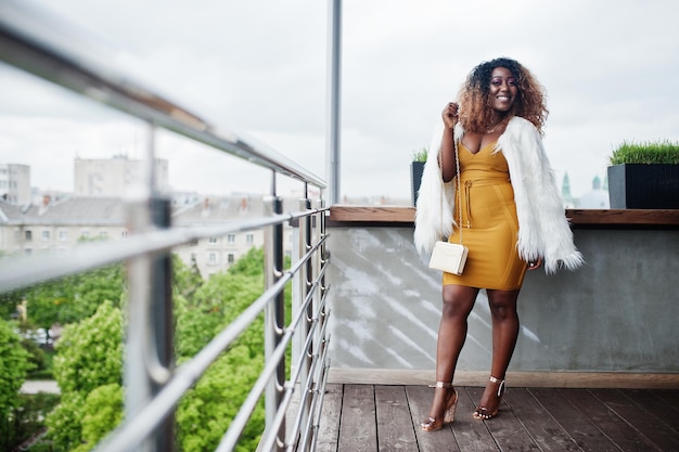Femme afro-américaine glamour en robe jaune et cape en laine blanche avec sac à main posé au balcon