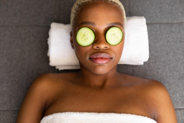 Photo une femme afro-américaine heureuse allongée sur une serviette avec des tranches de concombre sur les yeux. spa, relaxation, beauté et concept de style de vie.